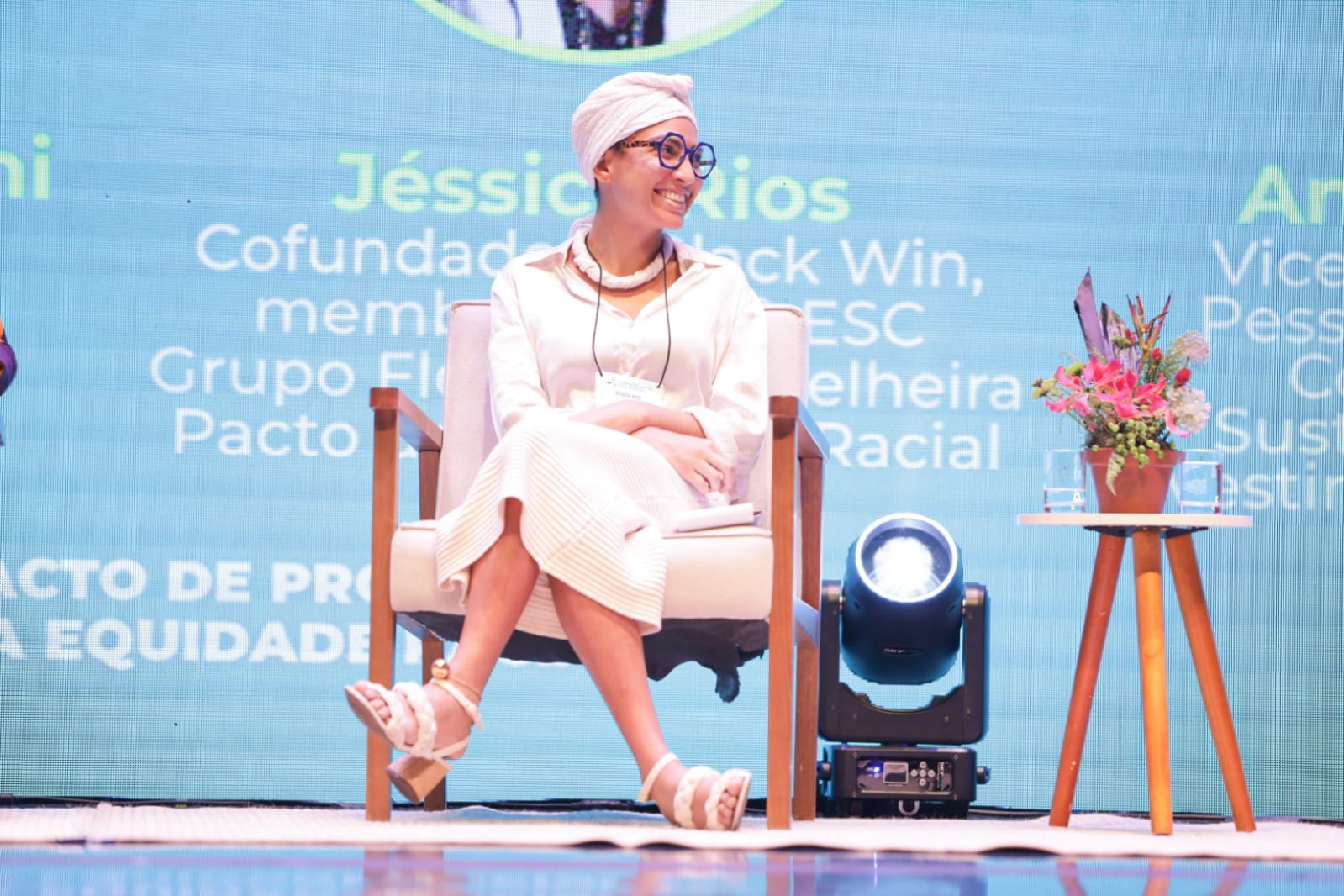 Jéssica Rios é uma mulher preta. Ela usa um turbante, colar, camisa de manga longa, saia e sandálias, todos os itens na cor branca em referência ao Candomblé. Está sentada em uma cadeira de madeira e usa óculos de aro preto.