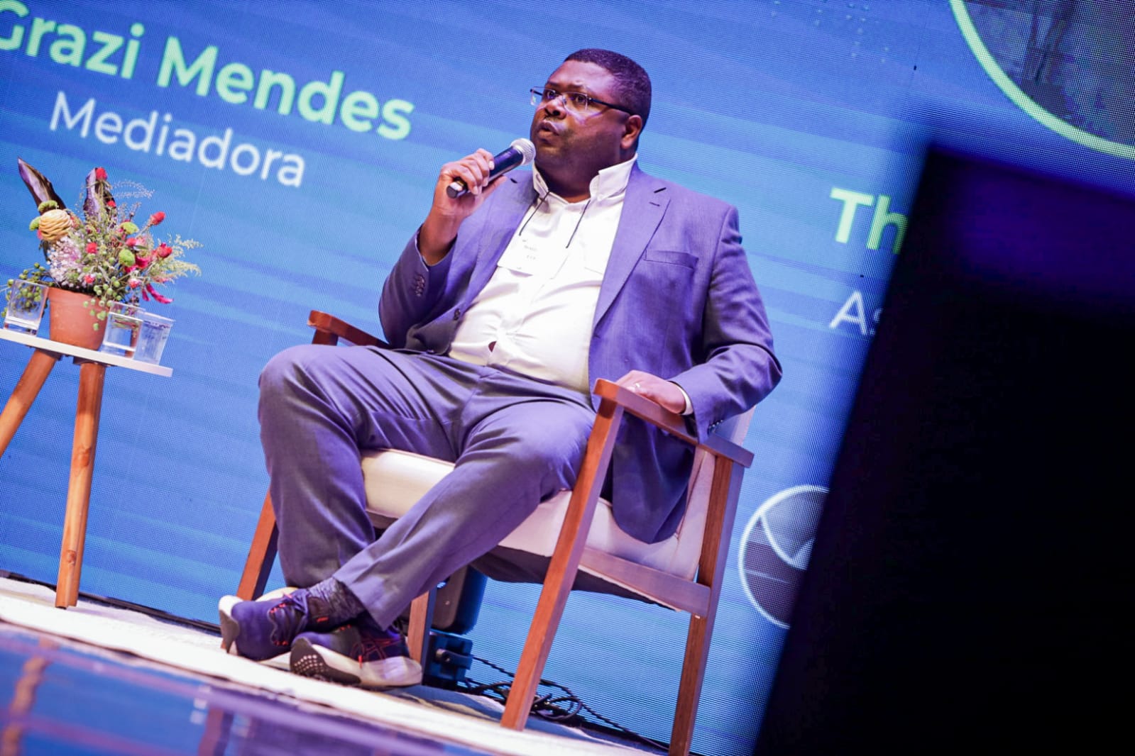Thiago Thobias é um homem preto, de cabelos pretos curtos. Veste camisa social branca com um terno azul e um tênis na mesma cor. Está sentado em uma cadeira de madeira e segura um microfone perto do rosto.