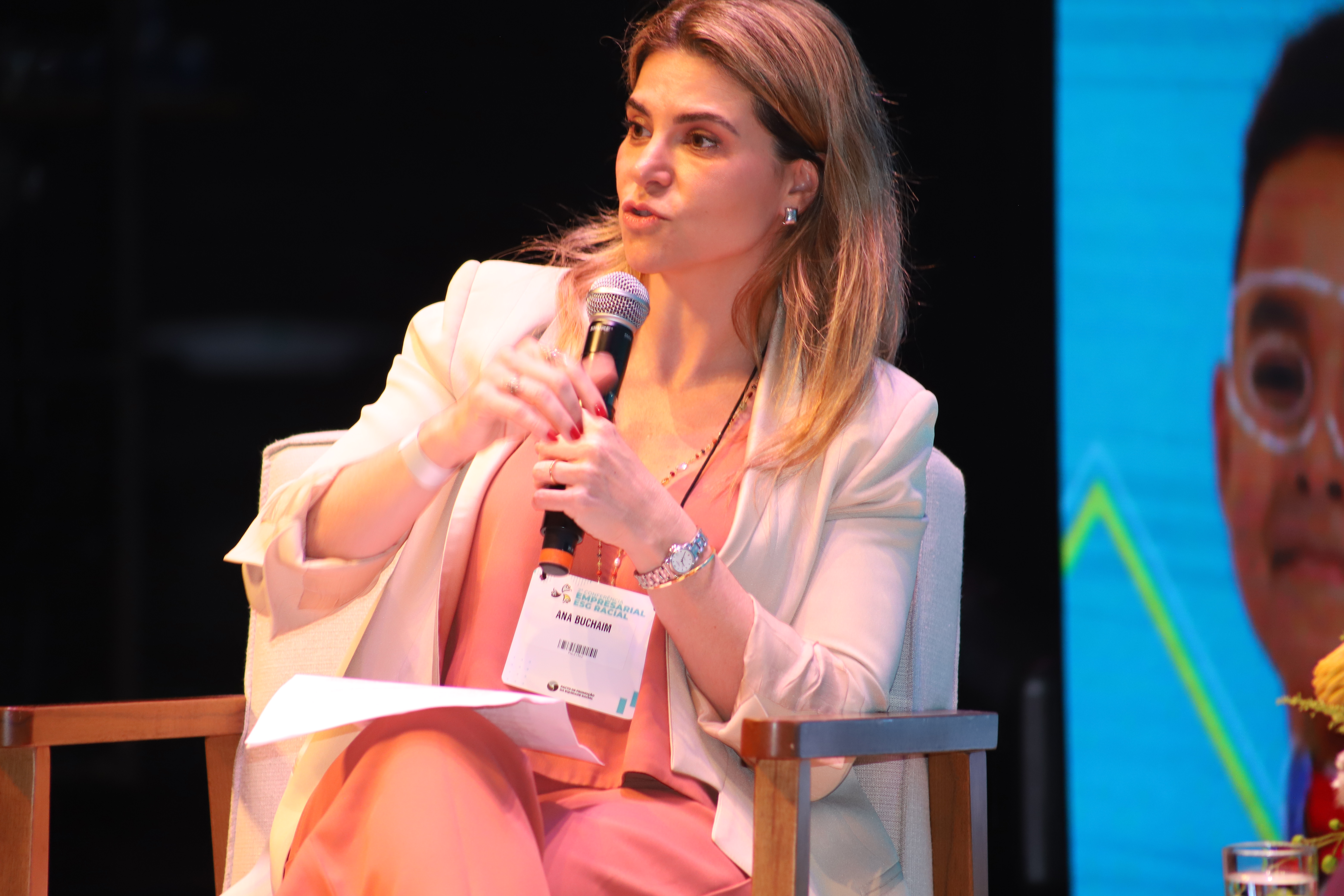 Ana Buchaim é uma mulher branca, de cabelos lisos loiros na altura do ombro. Veste um macacão coral e um terno branco. Está sentada em uma cadeira de madeira, segura um microfone perto do rosto e gesticula com a outra mão enquanto fala.